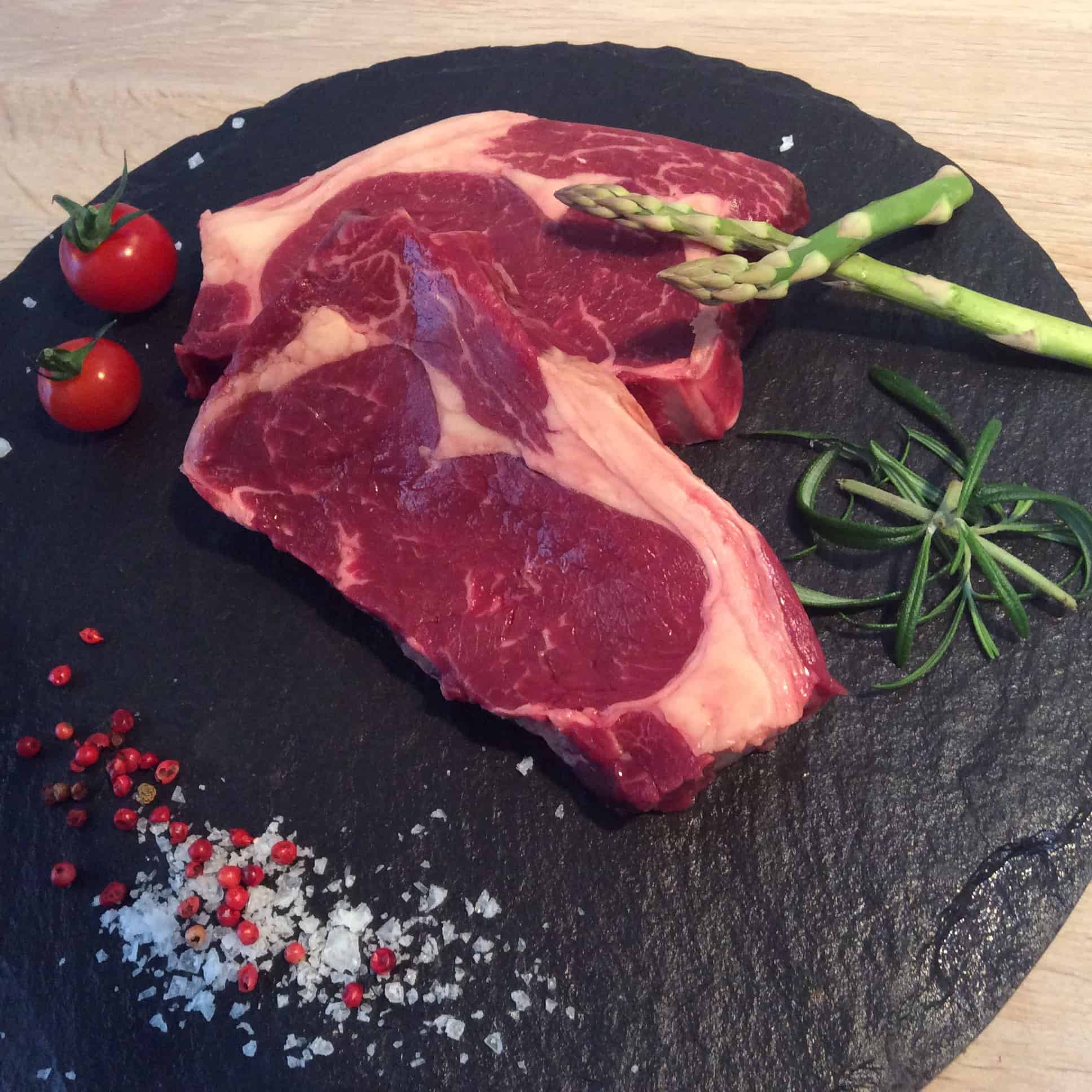 To rå ribeye-bøffer på en rund, sort skiferplade, pyntet med cherrytomater, asparges, rosmarin, peberkorn og havsalt, serveret på et træunderlag.