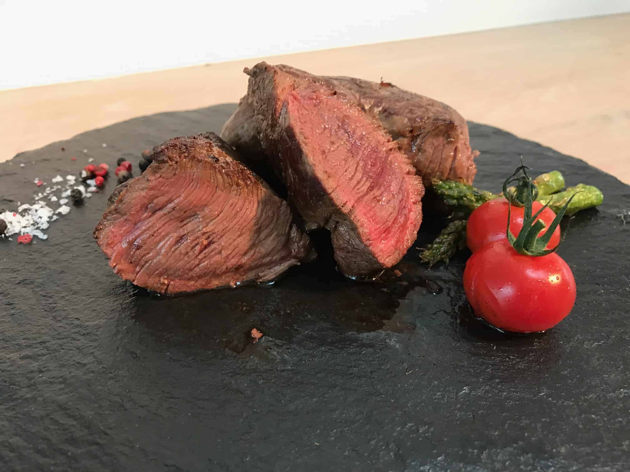 Tre skiver bøf, stegt medium-rare, er anrettet på en sort skiferplade sammen med asparges, cherrytomater og et drys salt og peber ved siden af.