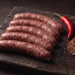 Fem rå bison grillpølser med chili på et mørkt træbræt, med en frisk chili og en grillet pølse ved siden af.