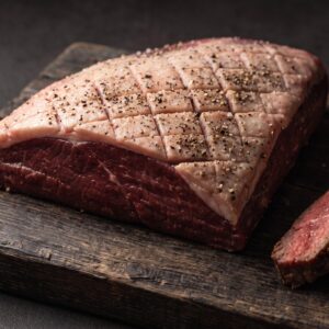 Rå culotte fra bison med ridset fedtkappe, salt og peber på et mørkt træbræt, med grillede skiver ved siden af.
