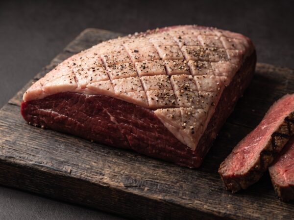 Rå culotte fra bison med ridset fedtkappe, salt og peber på et mørkt træbræt, med grillede skiver ved siden af.