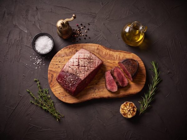Rå culotte fra bison med ridset fedtkappe på et oliventræsbræt, med grillede skiver ved siden af.