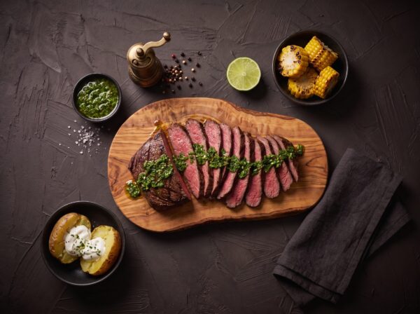 Tynde skiver grillet bison culotte med chimichurri på et bræt, med majs og bagte kartofler ved siden af.