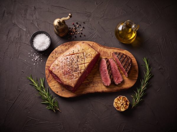 Rå culotte med ridset fedtkappe, salt og peber på et oliventræsbræt, med grillede skiver.
