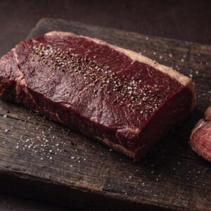 Rå filet af dansk bison med groft salt og peber på et mørkt træbræt, med stegte skiver med rød midte ved siden af.
