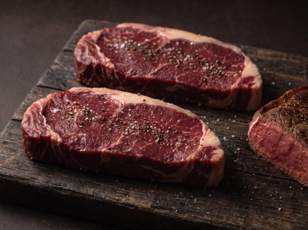 To rå bisonfilet-steaks med groft salt og peber på et mørkt træbræt, med en stegt steak med rød midte ved siden af.