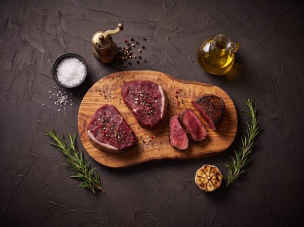To rå bisonfilet-steaks med salt og peber på et oliventræsbræt, med en stegt steak i skiver ved siden af.