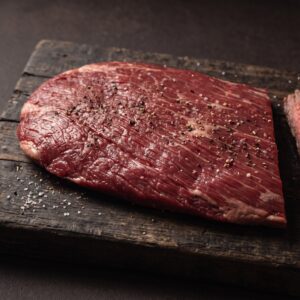 Rå flanksteak af skotsk højlandskvæg med tydelige fibre på et mørkt træbræt, med grillede skiver ved siden af.