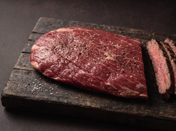 Rå flanksteak af skotsk højlandskvæg med tydelige fibre på et mørkt træbræt, med grillede skiver ved siden af.