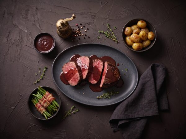 Skiver af stegt oksemørbrad med rødvinssauce på et fad, med kartofler og baconsvøbte bønner ved siden af.