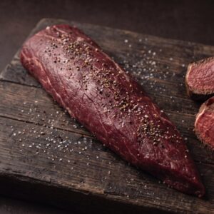Hel rå mørbrad fra bison med salt og peber på et mørkt træbræt, med stegte medaljoner ved siden af.