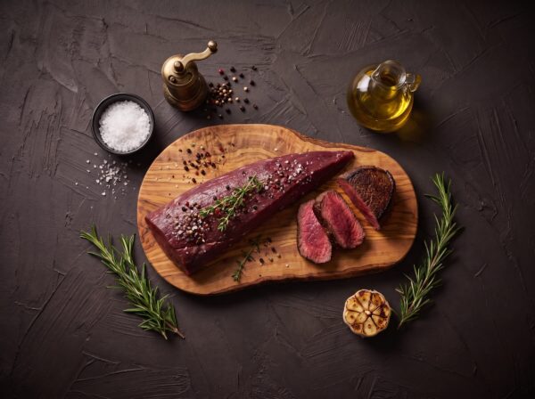 Hel rå mørbrad fra bison med salt, peber og timian på et oliventræsbræt, med stegte medaljoner ved siden af.