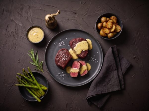 To medaljoner af bisonmørbrad med béarnaise på en mørk tallerken, med kartofler og grønne asparges ved siden af.