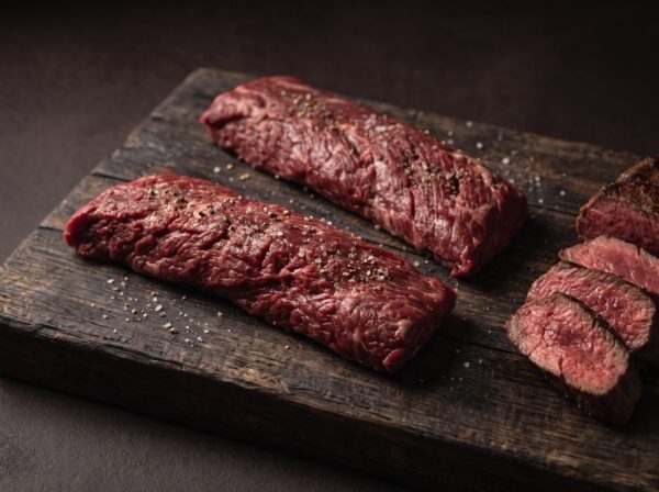 To rå nyretapper (onglet) af skotsk højlandskvæg på et mørkt træbræt, med stegte skiver ved siden af.