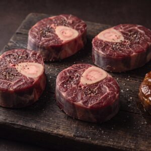 Rå osso buco af bison med marvben på et mørkt træbræt, med et braiseret stykke ved siden af.