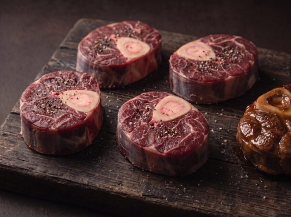 Rå osso buco af bison med marvben på et mørkt træbræt, med et braiseret stykke ved siden af.