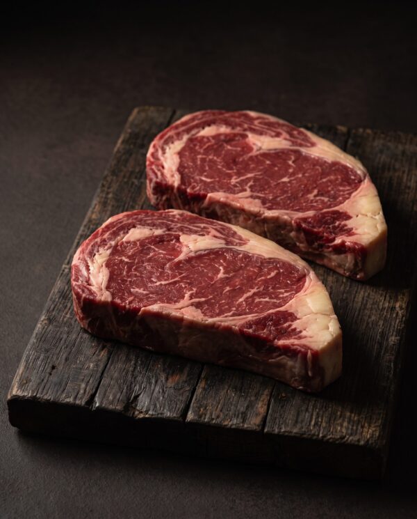 To rå ribeye-bøffer af skotsk højlandskvæg med fin marmorering og fedtkant, på et mørkt træbræt.