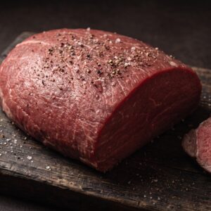 Hel rå rump heart filet af skotsk højlandskvæg på et mørkt træbræt, med stegte skiver ved siden af.