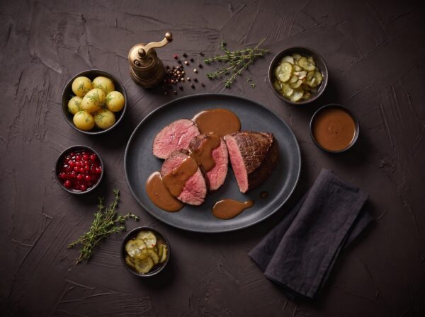 Skiver af stegt rump heart med brun flødesauce, med kartofler, ribsgelé og agurkesalat ved siden af.