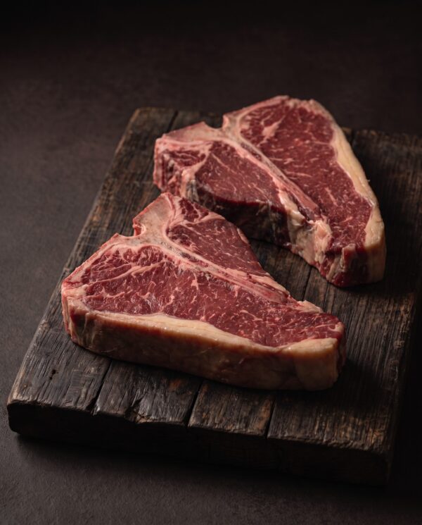 To rå T-bone steaks af skotsk højlandskvæg med det T-formede ben mellem filet og mørbrad, på et mørkt træbræt.