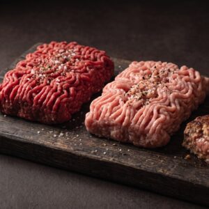 Hakket oksekød og hakket grisekød side om side på et mørkt træbræt, med stegte frikadeller ved siden af.