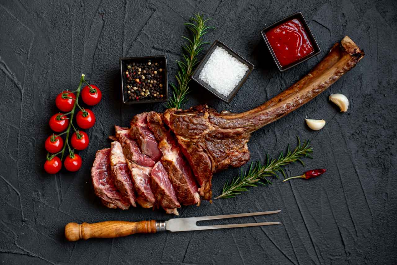 Skåret tomahawk-bøf med en stor knogle, omgivet af cherrytomater, rosmarin, hvidløg, chilipeber, salt, peberkorn og en skål med rød sauce på en mørk, struktureret overflade.